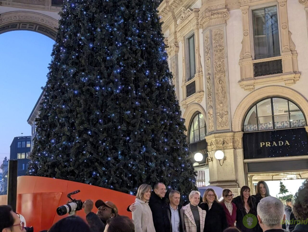 Albero di Natale decorato con luci LED blu in una piazza urbana storica. Albero di Natale decorato con luci blu a Milano, in un evento natalizio con un grande gruppo di persone, sotto un monumentale edificio storico con negozi di alta moda.