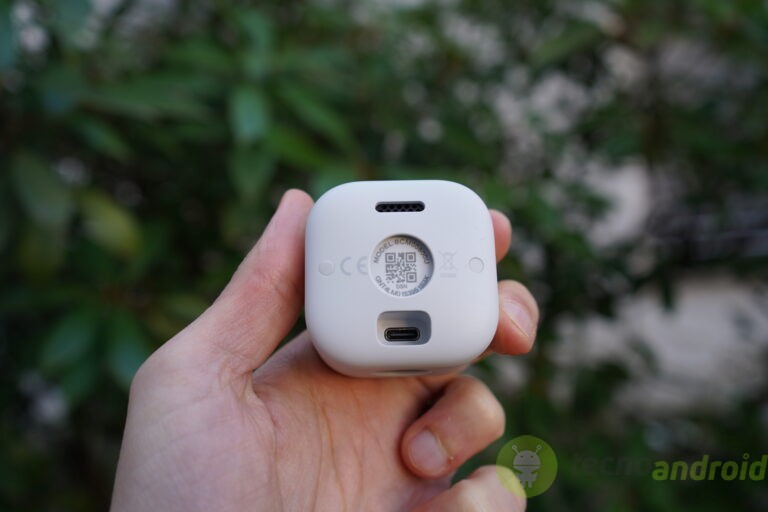 blink - immagine 37 Immagine di un auricolare Bluetooth bianco tenuto tra le mani, con evidenza sul retro, con QR code e dettagli tecnici, ambientato in uno spazio naturale con sfondo di foglie verdi.