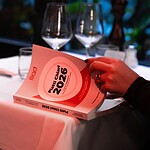 Alt text: Immagine di una persona che tiene in mano un libro bianco dal titolo "Piatta Chiari 2026" in un ristorante, con tavolo apparecchiato con bicchieri e posate, ambientazione elegante e moderna.
