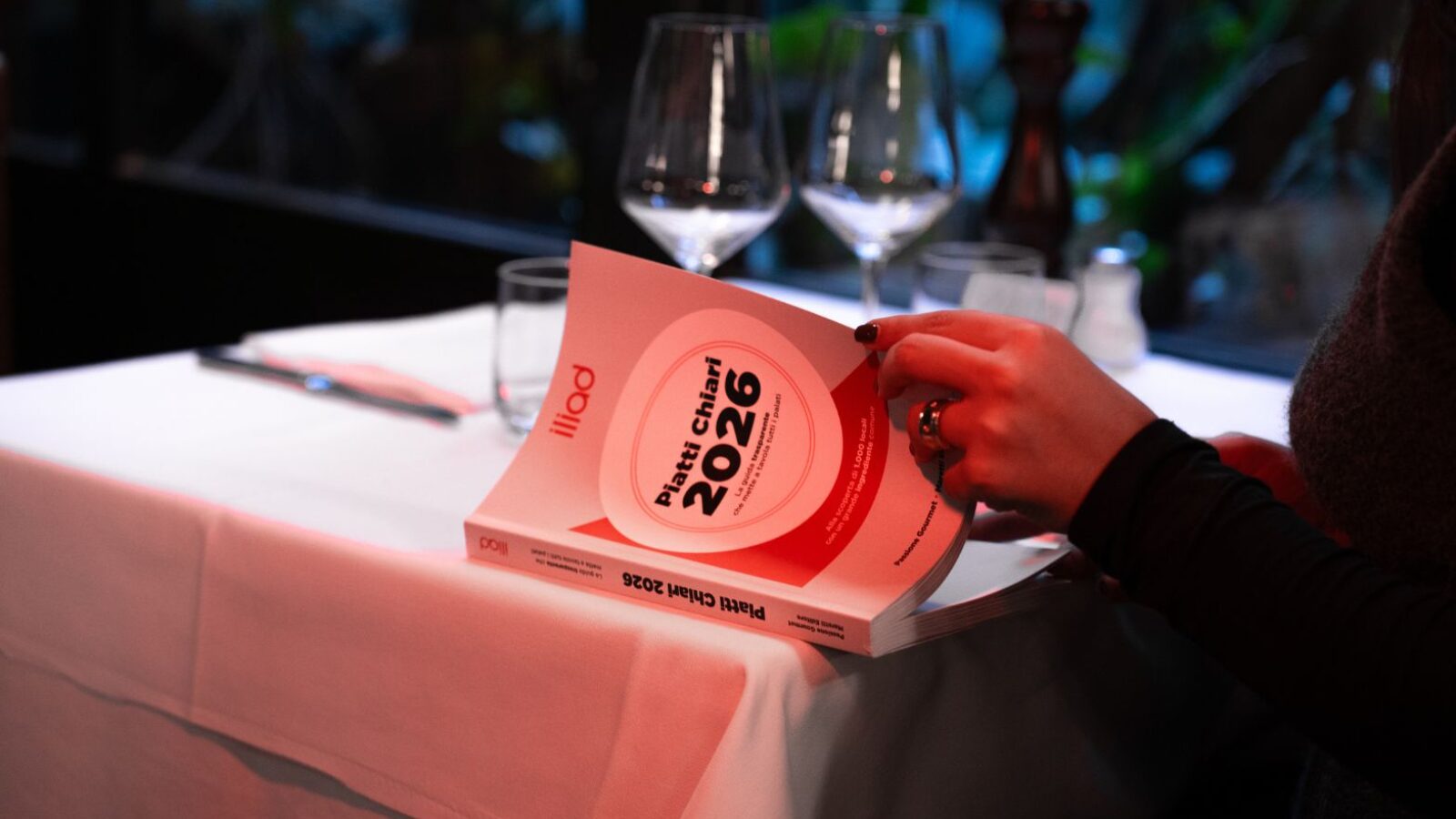 Titolo: Lettura Menù. Alt text: Immagine di una persona che tiene in mano un libro bianco dal titolo "Piatta Chiari 2026" in un ristorante, con tavolo apparecchiato con bicchieri e posate, ambientazione elegante e moderna.