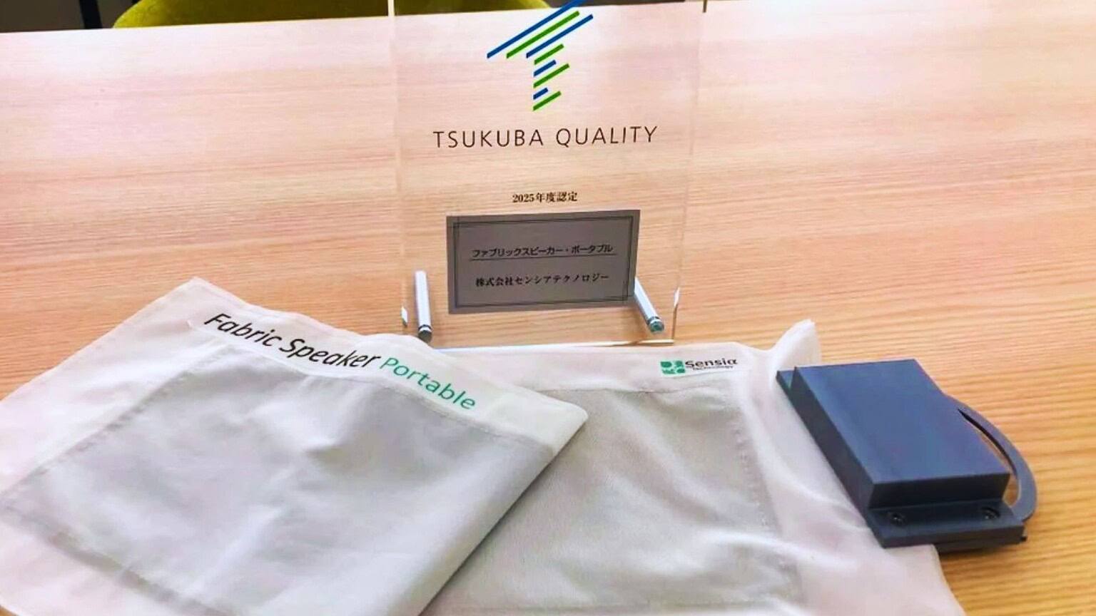 Altro: Immagine di un dispositivo portatile per altoparlanti tessili con logo Sensia, in esposizione su un tavolo in un'ambientazione professionale, promuovendo tecnologia audio da viaggio.
