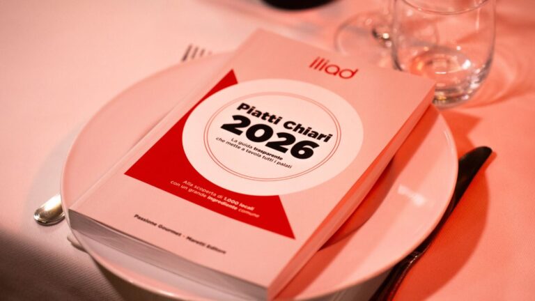 Alt text: Fotografia di un menu "Piatti Chiari 2026" posizionato su un piatto bianco, accompagnato da bicchieri d'acqua e posate su una tovaglia rosa, in un ambiente elegante.