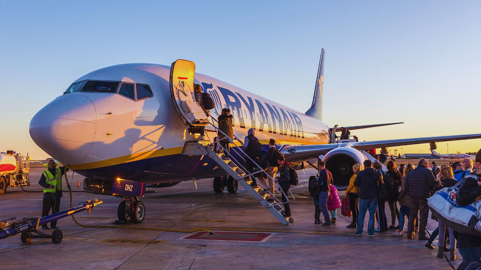 Immagine che mostra un aereo Ryanair parcheggiato con passeggeri in fila per salire, al tramonto, su un aereo low cost in partenza o in arrivo, in un aeroporto internazionale.