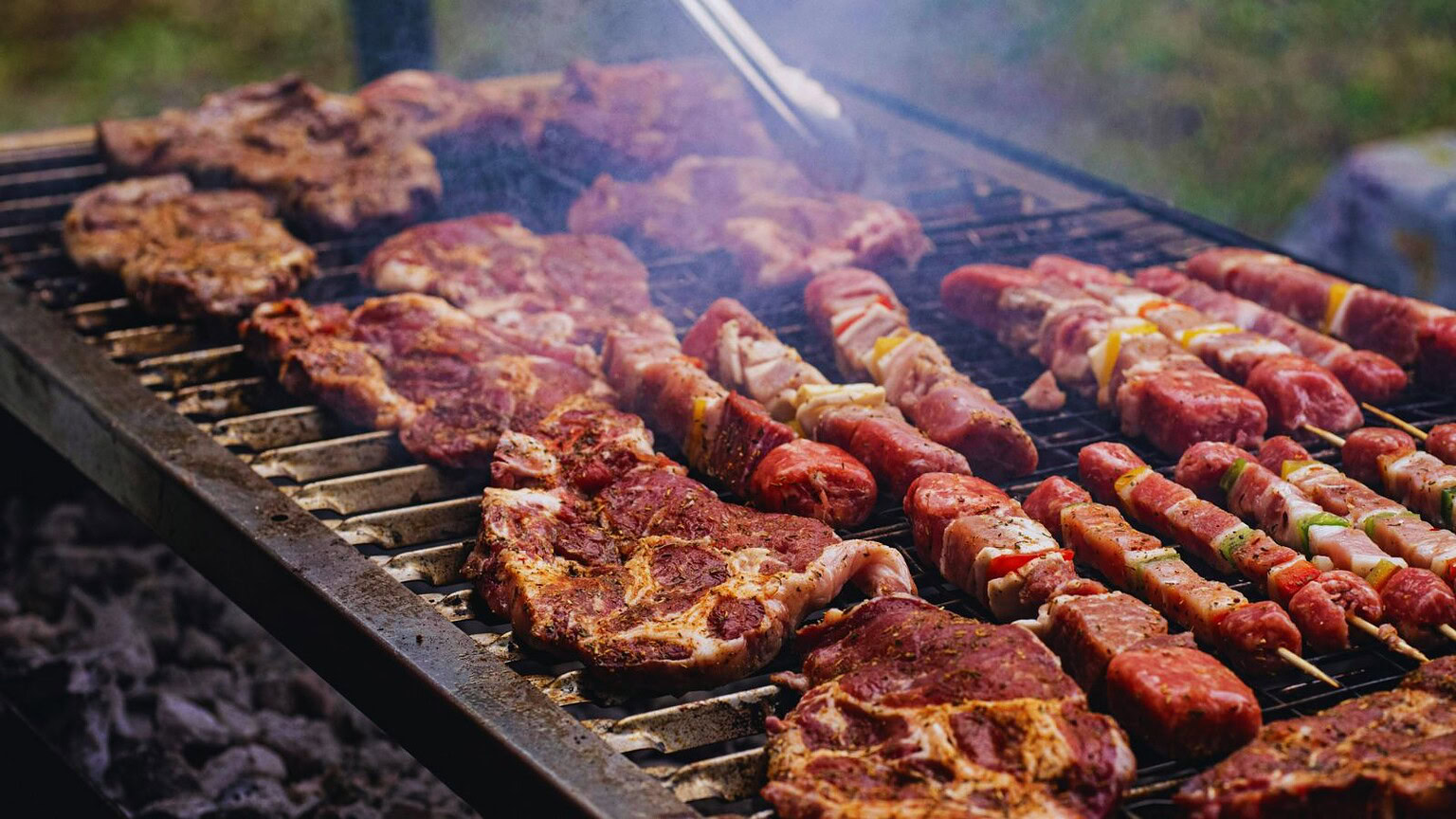 Carne e salsicce grigliate su barbecue, perfette per una festa o un picnic all'aperto, con fumo che sale e ambiente naturale di sfondo.