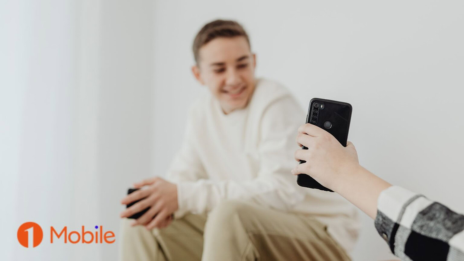 ALT text: Giovane uomo sorridente con smartphone nero, seduto in un ambiente chiaro, evidenziando l'uso di dispositivi mobili moderni.