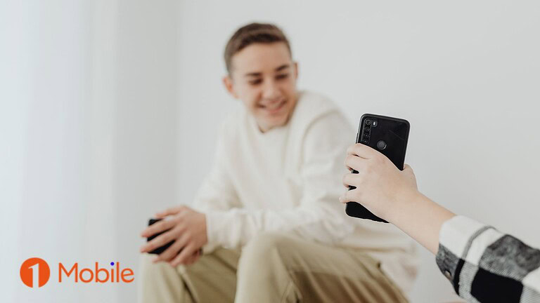 ALT text: Giovane uomo sorridente con smartphone nero, seduto in un ambiente chiaro, evidenziando l'uso di dispositivi mobili moderni.