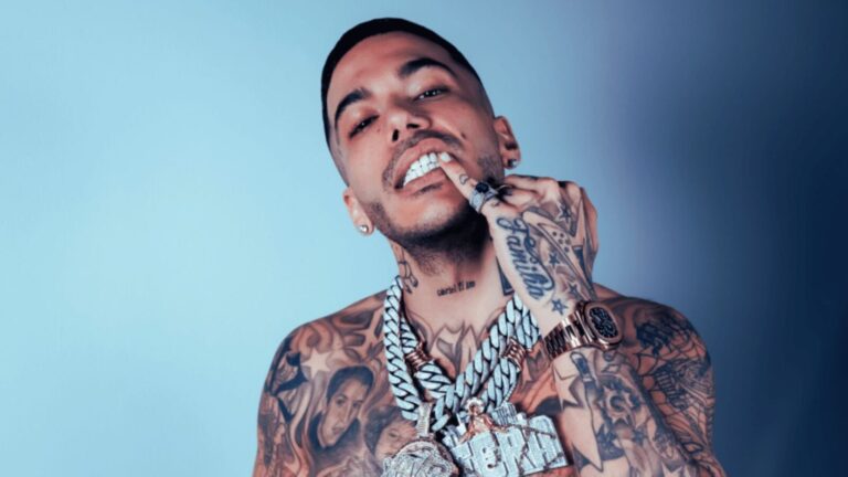 Rapper tatuato con gioielli di lusso, bracciali e collane d'oro, posa provocante su sfondo blu, stile streetwear appassionato, rappresentazione della cultura urbana e musica trap.