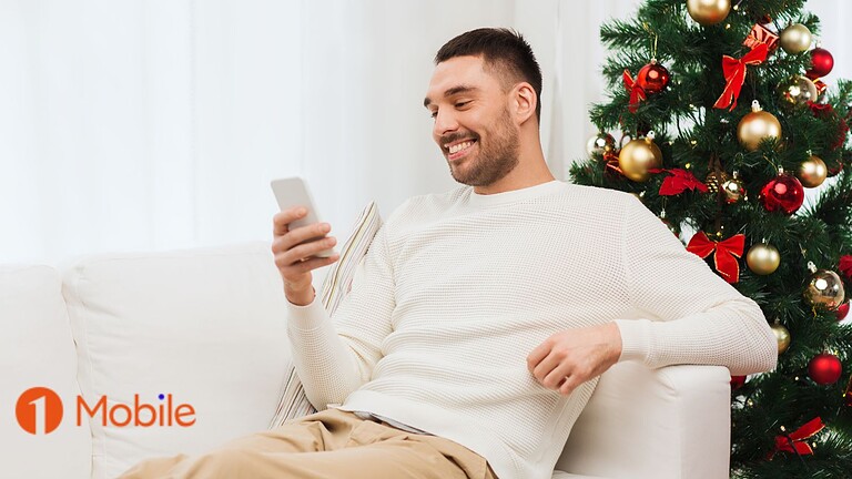 Immagine di un uomo che sorride utilizzando uno smartphone vicino a un albero di Natale decorato, rappresenta tecnologia, divertimento, festività e comunicazione digitale.