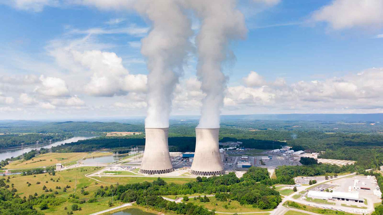 Immagine di due centrali nucleari con emettitori di fumo in un paesaggio naturale ricco di vegetazione e cielo nuvoloso, rappresentando l'energia nucleare e le centrali di produzione energetica sostenibile.